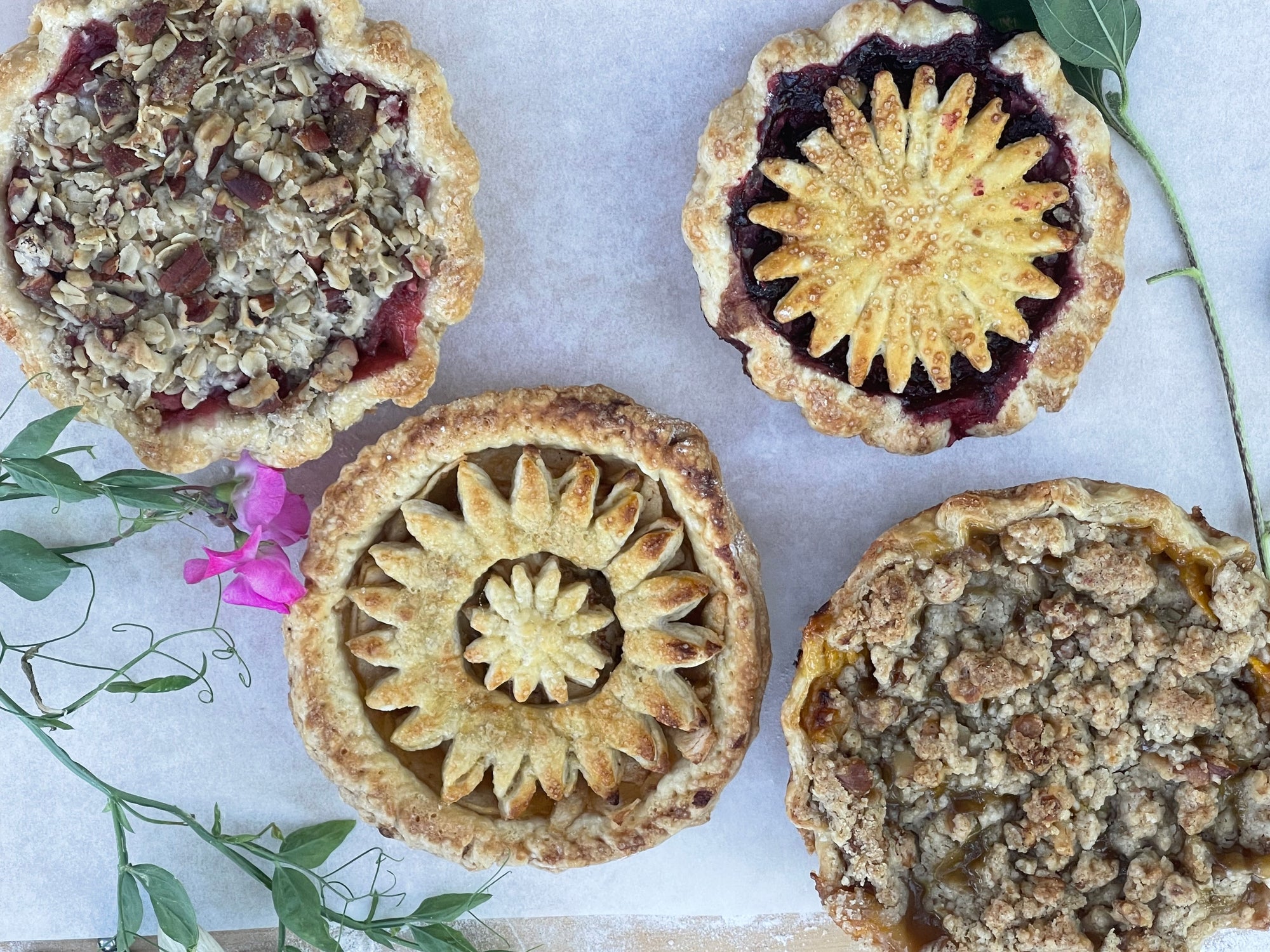 Four small fruit pies with different crust designs on a marble surface with greenery.